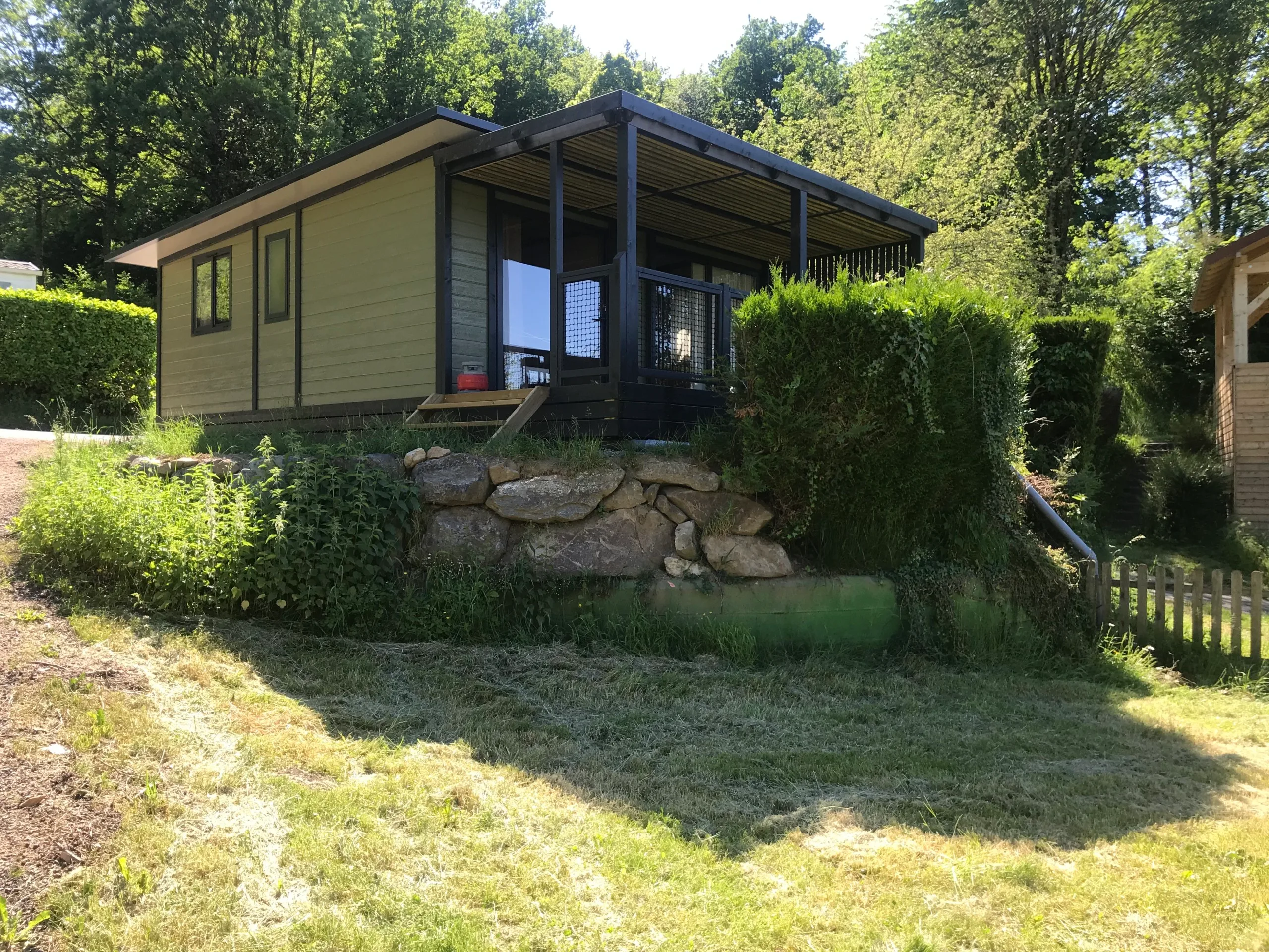 Chalet vert et gris avec terrasse couverte, situé sur un emplacement en pente avec muret en pierre naturelle, entouré d'arbres et de haies